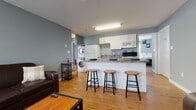 This single-floor unit offers a practical and well-organized layout across its main living areas. The open-concept living room and kitchen form the heart of the space, finished with warm hardwood-style flooring and cool grey walls throughout. The kitchen is equipped with white cabinetry, granite-style countertops, a full appliance suite including a range and built-in microwave, and a peninsula with bar seating for three — ideal for casual dining or entertaining. A stacked washer and dryer unit is conveniently integrated into the kitchen area, adding in-unit laundry access. The living room features large windows that admit generous natural light, a wall-mounted HVAC unit, and ample space for seating.

The unit includes two bedrooms, each carpeted for comfort. The primary bedroom offers dual closets and a connected en-suite bathroom with a glass-enclosed corner shower and white vanity. The second bedroom features a ceiling fan, a window with natural light, and its own HVAC unit. A second full bathroom serves this area, complete with a shower-tub combination and a white vanity. The consistent grey-toned walls and clean white trim throughout the unit create a cohesive, understated aesthetic well-suited to modern living.