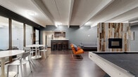 Located at 8000 E Girard Ave in Denver, CO, this single-floor media center and community lounge offers a well-appointed, open-concept layout designed for both relaxation and social interaction. The space features rich, dark hardwood-style flooring throughout, complemented by cool gray walls and exposed beam ceilings that lend an industrial yet welcoming character.

The main lounge area is thoughtfully arranged with vibrant orange accent chairs, a gray sectional sofa, and a full-size pool table as a central recreational focal point. A striking reclaimed wood accent pillar adds a rustic design element, anchoring the seating zone. A glass-walled dining area with multiple white round tables and modern chairs provides a bright, semi-private space ideal for gatherings or casual dining.

The kitchen features dark espresso cabinetry, white countertops, a stainless steel sink, dishwasher, and a full-size refrigerator, along with a peninsula bar with bar stools — perfect for casual entertaining. A hallway connects the various zones of the space and provides access to additional building amenities. Flush-mount ceiling lighting ensures consistent illumination throughout, making this media center a versatile and inviting communal destination.