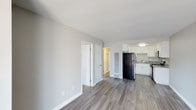 Unit #18 at 5865 El Cajon in San Diego is a single-floor residence that presents a clean, modern aesthetic throughout its open layout. The living area is finished with wood-style plank flooring in a warm gray tone, complemented by crisp white baseboards and smooth, light gray walls that lend the space a bright and airy feel. The open sightlines connect the main living area directly to the kitchen, creating a cohesive flow ideal for everyday living.

The kitchen is outfitted with white cabinetry, a stainless steel sink, and a black refrigerator, offering a functional and visually balanced workspace. The countertops appear to feature a speckled granite-style finish, adding a subtle detail to the overall design. Recessed and flush-mount lighting ensures the kitchen remains well-lit.

A hallway leads from the main living space to additional rooms, with a set of bifold closet doors visible along the wall, providing convenient storage. A frosted glass panel element adds a subtle design accent within the corridor. The overall ambiance is clean and move-in ready, reflecting a straightforward and practical living environment in San Diego's El Cajon Boulevard corridor.