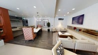 This single-floor space is thoughtfully designed to accommodate both dining and lounging, offering a versatile and welcoming environment. The layout is divided into two well-defined zones that flow seamlessly into one another.

The dining area features a long white rectangular table surrounded by striking high-back chairs upholstered in a bold coral and white chevron pattern, creating a vibrant focal point. Above the table, a modern branching chandelier adds an artistic touch, complementing the recessed lighting throughout the ceiling.

Adjacent to the dining area, the lounge zone is furnished with a collection of neutral-toned armchairs and a low coffee table arranged atop a geometric-patterned area rug. A floor lamp provides warm ambient lighting, while a wall-mounted television and a minimalist ledge with decorative accents contribute to the room's polished aesthetic.

Dark wood-look tile flooring spans the entire space, grounding the contemporary design and providing a cohesive visual foundation. A potted indoor tree adds a natural element, and framed artwork on the walls enhances the curated atmosphere. The space is well-lit and offers a refined, modern ambiance suitable for both social gatherings and relaxed activities.