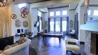 This elegantly designed single-floor space functions as a upscale communal lounge or clubhouse, immediately impressing with its soaring double-height ceilings and expansive floor-to-ceiling windows dressed in rich, dark curtains. Clerestory windows positioned above the main window line flood the interior with abundant natural light, creating a bright and welcoming atmosphere throughout.

The flooring features a striking herringbone-patterned hardwood, adding a refined architectural element that anchors the room's sophisticated aesthetic. A full-size billiards table serves as the central focal point, complemented by an eclectic mix of seating including plush cream-toned sofas and contemporary accent chairs.

Distinctive decorative lighting includes an ornate, sculptural chandelier alongside a classic pendant fixture, contributing to the room's layered and curated ambiance. A wall-mounted flat-screen television and a dedicated computer workstation area enhance the space's functionality.

Tall potted plants are strategically placed throughout, introducing organic warmth and texture. Subtle crown molding and coffered ceiling details further reinforce the refined, upscale character of this well-appointed gathering space.