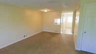 This two-floor home offers a straightforward and functional layout suited for everyday living. The main living area is finished with wall-to-wall carpet in a warm neutral tone, providing a comfortable and inviting atmosphere. Smooth, light-colored walls throughout the space contribute to a clean and airy feel, while flush-mount ceiling lighting ensures consistent illumination. A textured ceiling adds a subtle architectural detail to the room. Ventilation registers are thoughtfully integrated into both the floor and walls, indicating a central HVAC system for year-round climate control. A pass-through window connects interior spaces, enhancing the open flow between rooms. The entryway, visible from the main living area, features hard-surface flooring and benefits from natural light streaming through a glazed front door, creating a welcoming point of entry. White trim and baseboards provide clean contrast against the neutral wall tones, lending a polished finish to the interior. The overall design is practical and understated, with a neutral palette that offers flexibility for a variety of furnishing and décor styles. Spanning two floors, the home provides ample space for both living and private areas.