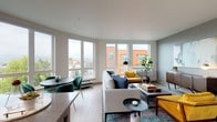 This single-floor residence showcases a contemporary design with an open, flowing layout connecting the main living spaces. The living room is a standout feature, bathed in abundant natural light through a generous wrap-around bank of floor-to-ceiling windows that frame sweeping urban views of the surrounding cityscape. Light wood-tone flooring runs throughout, complementing the neutral color palette accented by warm, modern furnishings.

The kitchen is thoughtfully designed with rich wood-grain cabinetry, stainless steel appliances including a refrigerator, range, and built-in microwave, and a sleek gray quartz countertop. A peninsula island provides additional workspace and casual seating. A geometric-patterned tile backsplash adds a refined decorative touch.

The dining area sits adjacent to the living room, benefiting from the same expansive windows and natural light. Two well-proportioned bedrooms offer a calm retreat; one includes a walk-in closet, while both feature light wood flooring and neutral walls. The bathrooms are finished with wood-tone vanities, gray quartz countertops, large mirrors, and glass-enclosed showers, delivering a clean, modern aesthetic throughout.