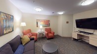 This single-floor library is thoughtfully designed to serve multiple functions, combining a relaxed lounge environment with practical meeting and study areas. The space is unified by patterned carpeting and flush-mount ceiling lighting that provides consistent, even illumination throughout.

The lounge area is furnished with a dark upholstered sofa, plush orange armchairs, and a round coffee table, creating an inviting setting for casual reading or conversation. Colorful framed artwork lines the walls, adding vibrancy to the neutral palette. A floor lamp and table lamp supplement the overhead lighting, contributing to a warm ambiance.

Adjacent to the lounge, a bold red accent wall defines the reading and meeting zone, where freestanding bookshelves are stocked with a diverse collection of titles. A light wood oval conference table surrounded by black task chairs provides ample space for group discussions or collaborative work. A complementary piece of artwork decorates this area as well.

A dedicated workstation with a wall-mounted flat-screen television and a light wood desk offers additional functionality. The entry features a solid wood door with a sidelight panel, providing a clear and accessible point of entry to the space.