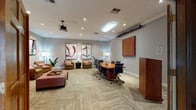 This well-appointed single-floor professional space blends classic architectural character with modern functionality. The entrance area is defined by striking floor-to-ceiling wood columns with paneled detailing, flanking a built-in workstation wall outfitted with dual computer monitors, shelving, and ample desk surface — ideal for focused individual work. Beyond the columns, a bright lounge area features a set of wingback and accent chairs arranged around a low coffee table, bathed in natural light from a prominent arched French door entry that lends warmth and openness to the space.

A central multi-purpose zone seamlessly connects the lounge with a formal conference setup, where a polished wood conference table is surrounded by ergonomic task chairs. The room is equipped with a ceiling-mounted projector, wall-mounted display panel, and a presentation lectern, making it well-suited for meetings and presentations. Recessed lighting and crown molding throughout contribute to the polished aesthetic.

The lounge seating areas are furnished with tufted leather chairs accented with pink cushions, complemented by dark side tables and contemporary floor lamps. Bold abstract artwork adorns the neutral gray walls, adding visual interest. Herringbone-patterned carpet flooring ties the zones together, creating a cohesive and professional yet inviting atmosphere throughout.