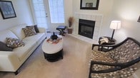This single-floor space offers a thoughtfully designed living area that balances comfort and style. The room centers around a classic white-tiled fireplace with a dark surround and a framed mirror above the mantle, serving as a striking focal point. Plush, neutral-toned carpeting runs throughout, lending warmth and a soft underfoot feel to the space.

Natural light pours in through large windows fitted with horizontal blinds, brightening the interior and creating an airy, open ambiance. The layout comfortably accommodates a diverse seating arrangement, including a sleek white sofa adorned with decorative accent pillows, patterned upholstered chairs, and a stylish oval coffee table with a tiered base, all contributing to a cohesive and inviting aesthetic.

Decorative touches such as a tall dried floral arrangement and tasteful tabletop accessories add character without overwhelming the room. A glass-paned exterior door provides an additional source of light and easy access to the outdoors. The overall design palette leans toward a neutral scheme of whites, beiges, and dark accents, resulting in a clean, elegant atmosphere that is both functional and visually appealing.