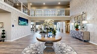 Located on 25th Ave NE in Aberdeen, South Dakota, this elegantly designed community space makes a striking first impression with its soaring two-story lobby. The grand entrance is anchored by a circular wooden centerpiece table adorned with lush floral arrangements, set atop a decorative area rug, and framed by textured feature walls with a soft, neutral palette. Dramatic double-height ceilings, a statement chandelier, and an ornate wrought-iron upper-level balustrade lend the space a refined, upscale character.

The open layout flows seamlessly from the lobby into a generously sized dining and gathering area, bathed in natural light from a row of tall windows along a contrasting charcoal accent wall. This area comfortably accommodates multiple dining tables and chairs, creating an inviting atmosphere for communal meals and events. The wood-look flooring throughout ties each zone together cohesively.

A well-equipped kitchen area features white cabinetry, a large stainless steel refrigerator, an island with bar seating, and patterned accent walls, offering full functionality for events or daily service. The private office is neatly furnished with a substantial desk, filing storage, and wall art, providing a quiet and productive workspace. A connecting hallway with curated artwork and seating nooks completes the thoughtfully designed interior.