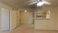 This single-floor residence offers an open-concept layout that seamlessly connects the living area and kitchen, creating a sense of spaciousness and ease of movement. The living space is finished with neutral beige carpeting and light-colored walls, lending a warm and understated ambiance throughout. A ceiling fan with an integrated light fixture serves as a functional centerpiece overhead, providing both air circulation and illumination to the main living area. The kitchen is positioned adjacent to the living space and features a raised breakfast bar counter, natural wood-toned cabinetry, and a built-in range with an overhead ventilation hood. This open pass-through design between the kitchen and living area encourages an interactive and connected feel. A utility area near the entryway includes a louvered storage closet and what appears to be a built-in desk or countertop nook, adding practical storage and workspace options. Recessed and flush-mount ceiling lighting provides consistent illumination throughout. The overall aesthetic is clean, neutral, and functional, making it a versatile space well-suited for comfortable everyday living.