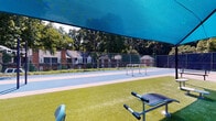 Located at 150 Hunt Club Lane in Raleigh, NC, this outdoor recreational space offers a well-equipped amenity area designed for both sport and fitness. The centerpiece of the space is a set of hard-surface courts finished in blue and gray, lined for tennis and pickleball and enclosed by chain-link fencing with privacy screening along the perimeter. A small goal frame is positioned on the court surface, adding versatility to the space.

Adjacent to the courts is a dedicated fitness and relaxation zone surfaced with lush artificial turf. This area is sheltered by a large, vibrant blue shade canopy supported by sturdy posts, providing relief from direct sunlight during outdoor workouts. Several outdoor fitness stations are distributed across the turf, including a dual-rider cycling machine, a stretching and ab station, and additional resistance-based equipment, catering to a range of exercise preferences. A park-style bench is also available for rest or spectating.

The entire area is framed by mature trees that provide a natural backdrop and additional shade, contributing to a comfortable, park-like atmosphere. Brick residential buildings are visible beyond the fencing, situating this recreational space within a broader community setting.