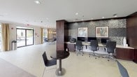 This internet café is a well-appointed, single-floor facility that seamlessly blends a functional business center with comfortable communal areas. The workstation zone is defined by a sleek, built-in desk surface accommodating multiple computer terminals, a multifunction printer, and ergonomic task chairs. A striking mosaic tile accent wall in shades of silver and charcoal serves as a dramatic backdrop, complemented by framed contemporary artwork that adds a refined visual element. Recessed lighting throughout ensures consistent illumination across all work areas.

Adjacent to the business center, an open lounge area provides a welcoming retreat with light-toned tile flooring transitioning into carpeted seating zones. Clustered seating arrangements feature upholstered chairs in muted yellows and greens centered around low tables, while a chandelier and large windows with draped curtains contribute natural light and an airy atmosphere. Bistro-style tables offer additional casual seating throughout the shared space.

Frosted glass sliding doors lead to transitional corridors, and dark wood column accents provide architectural contrast against the predominantly white walls. Framed artwork is thoughtfully placed throughout, reinforcing a cohesive, polished aesthetic suitable for both focused work and relaxed social interaction.