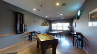 This single-floor game room is designed for entertainment and socializing, combining a polished aesthetic with a variety of recreational amenities. The space is anchored by rich hardwood flooring that runs throughout, complementing the deep blue-gray walls that lend the room a moody, sophisticated atmosphere.

At the center of the room, a classic foosball table takes pride of place, offering an inviting focal point for guests. Just beyond it, a full-size pool table is positioned beneath a striking set of antique-style brass pendant lights suspended by chains — a standout design element that adds vintage character to the space. A wall-mounted cue rack keeps equipment neatly organized nearby.

Natural light filters in through a pair of windows along the far wall, brightening the interior and balancing the warm glow of recessed ceiling lights. An arcade machine is tucked to the side, adding a retro gaming element to the room's entertainment offerings. Bar-height seating and a tall table provide comfortable spots to relax between games.

The overall ambiance is that of a well-appointed, thoughtfully curated recreation space — equal parts functional and stylish, ideal for casual gatherings and friendly competition alike.