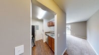 Located at 624 Wallen Hills Dr in Fort Wayne, Indiana, this single-floor residence offers a practical and well-organized layout with one bedroom and two bathrooms. The entrance leads into a hardwood-style floored kitchen, which features warm wood cabinetry, dark granite-style countertops, a sink, dishwasher, and full-size refrigerator. A pass-through window connects the kitchen to the carpeted living room, creating an open sightline between the two spaces. The living room is generously proportioned relative to the overall footprint, bathed in natural light from a large sliding glass door that opens to an outdoor patio with views of the surrounding landscaping. Neutral gray walls and plush carpet throughout the living and bedroom areas lend a clean, cohesive feel. The bedroom is a comfortable, carpeted space with a window providing natural light, and it connects directly to one of the two bathrooms, which includes warm wood vanity cabinetry and a mirrored medicine cabinet. The second bathroom features tile flooring and a tub-shower combination. Additional utility is provided by a dedicated laundry area, utility room, and two closets, maximizing storage within the compact footprint.