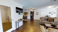 This single-floor space showcases a comfortable, open-concept layout that allows the living, dining, and entry areas to flow naturally into one another. Rich, dark hardwood-style flooring runs throughout, anchoring the space with a warm, cohesive feel that contrasts cleanly against the bright white walls.

The living area is positioned to the right upon entry and features a grey leather sofa accented with decorative pillows, complemented by a wall-mounted flat-screen television on the adjacent wall. A sleek metal-framed console table beneath the TV provides a functional surface for décor, including ceramic accents and decorative vases.

Crown molding lines the ceiling throughout, adding a refined architectural detail, while a textured ceiling with a centrally placed ceiling fan and light fixture provides both style and practicality. The dining area, situated toward the rear of the open space, accommodates a compact dark wood table and matching chairs, ideal for everyday dining.

A dark wood bookcase near the back wall offers display and storage space, decorated with baskets and framed items. Framed wall art and subtle decorative touches throughout give the space a welcoming, well-appointed character.