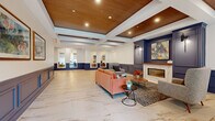 This generously sized single-floor space functions as a sophisticated lobby and corridor area, thoughtfully designed to make a strong first impression. The layout comprises an expansive central lounge and two connecting hallways, creating a seamless flow throughout the space.

The lounge area is anchored by a sleek linear fireplace set into a navy blue built-in surround, topped with a bold piece of contemporary artwork and flanked by decorative wall sconces. Comfortable seating arrangements, including a salmon-toned sofa, armchairs, and accent tables, create an inviting gathering environment. The standout ceiling treatment features coffered panels with warm wood insets and recessed lighting, adding architectural depth and visual warmth.

Underfoot, large-format marble-look porcelain tiles in cream and gold tones extend consistently throughout the lounge and hallways, contributing to a clean, upscale aesthetic. Navy blue wainscoting with raised panel detailing lines the walls throughout, providing a cohesive design language across all areas.

The hallways connect to elevator banks finished in brushed stainless steel and lead toward glass-panel entry doors, allowing natural light to filter through. Full-length mirrors are integrated into hallway walls, enhancing the sense of openness and depth throughout this well-appointed space.