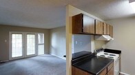 Located in Tyler, Texas, this single-floor residence offers a practical and comfortable layout designed for everyday living. The kitchen is outfitted with dark granite-style countertops, rich wood-toned upper cabinetry, and a white electric range, providing a functional cooking space. A half-wall partition separates the kitchen from the adjoining living area, creating an open-concept feel while maintaining a sense of definition between spaces. The living area features neutral carpet flooring and soft, warm wall tones that contribute to a welcoming ambiance throughout. A sliding glass door or large window at the far end of the living space invites natural light deep into the interior and offers a view of the lush greenery outside. The textured ceiling adds a subtle architectural detail consistent throughout the unit. The overall layout is straightforward and efficient, making it well-suited for those seeking a low-maintenance, single-level living arrangement in the Tyler, TX area.
