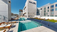 Located at 1025 N Park Ave in Tucson, Arizona, this rooftop amenity level at the Malibu Tucson community offers a well-appointed outdoor retreat designed for relaxation and socializing. The centerpiece of the deck is a resort-style swimming pool with crystal-clear water, complemented by an adjacent spa, both finished with clean, light-toned tile surrounds. Generous lounging areas flank the pool, featuring rows of cushioned wood-framed chaise lounges arranged on lush artificial turf and large-format porcelain tile, providing ample space for sunbathing beneath the abundant Tucson sunshine. A warm wood deck anchors additional gathering zones, including an outdoor kitchen equipped with a built-in grill, perfect for entertaining. The rooftop also features a glass-walled interior lounge or clubroom with floor-to-ceiling folding glass doors that open fully to the outdoor terrace, seamlessly blending indoor and outdoor living. The polished concrete and neutral-toned architectural finishes of the surrounding Malibu building lend a modern, contemporary aesthetic to the space. Panoramic views of the Tucson cityscape and the surrounding mountain ranges, including the Santa Catalinas, create a dramatic backdrop that enhances the open-air experience throughout the deck.