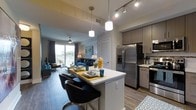 This single-floor residence showcases a thoughtfully designed open-concept layout that seamlessly connects the kitchen, dining, and living areas. The kitchen is outfitted with stainless steel appliances, including a French door refrigerator and gas range, complemented by medium-toned wood cabinetry and light-colored countertops. A central island with a white quartz-style surface doubles as a breakfast bar, accommodating seating for two with modern black chairs. Pendant lighting hangs above the island, while track lighting illuminates the kitchen workspace.

The open living area features wood-look flooring throughout, creating a warm and cohesive aesthetic. A ceiling fan keeps the space comfortable, and large sliding glass doors flood the room with natural light while providing direct access to an outdoor area with views of mature landscaping. Neutral wall tones are paired with contemporary furnishings, contributing to a clean and inviting ambiance. A closeted storage area is also visible, adding practical functionality. The overall design balances modern style with everyday livability.
