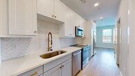 Located at 4312 Yancey Rd in Charlotte, NC, this thoughtfully designed single-floor unit makes excellent use of its compact footprint. The space is finished with light hardwood flooring throughout, crisp white walls, and clean modern trim, creating a bright and cohesive aesthetic.

The galley-style kitchen is a standout feature, showcasing white shaker upper cabinets paired with gray lower cabinets, all accented by brushed gold hardware. A herringbone-patterned white tile backsplash adds visual interest, while the white quartz countertops provide a sleek workspace. Stainless steel appliances, including a range, microwave, and dishwasher, complete the kitchen's functional design. A gold-toned industrial-style faucet over a deep undermount sink adds a refined touch.

The living area is open and airy, benefiting from a bank of large windows dressed with white blinds that invite generous natural light and offer views of the surrounding neighborhood. The clean, neutral walls provide a versatile backdrop for any furnishing style.

The bathroom features a wide gray vanity with ample drawer storage, a white undermount sink, quartz countertop, and a large mirror framed in matte black. Light wood-look flooring continues throughout.

A walk-in closet offers multi-level shelving and hanging rods along two walls, maximizing storage efficiency within the unit's overall design.