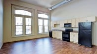 This single-floor space showcases a distinctive loft-inspired character, blending industrial and functional design elements throughout. The open-concept layout creates a generous sense of room, with rich, dark hardwood flooring extending across the entire main area, grounding the space with warmth and contrast.

A defining architectural feature is the row of large, arched-top windows along the front wall, which flood the interior with abundant natural light while offering direct views of the surrounding urban streetscape. The elevated ceiling height enhances the sense of openness, and exposed metal ductwork running along the ceiling adds an authentic industrial aesthetic.

The kitchen is positioned along one wall and is well-equipped with cream-toned cabinetry offering ample storage, dark laminate countertops, a black electric range, built-in microwave, and a double-basin sink. The kitchen integrates seamlessly into the open living area without visual obstruction.

Neutral wall tones throughout keep the space feeling clean and versatile. The combination of urban views, high ceilings, natural light, and industrial detailing makes this a characterful and functional living environment suited to a variety of uses.