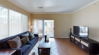 This single-story residence offers a practical and comfortable layout across several distinct living spaces. The living room serves as the heart of the home, featuring warm hardwood-style flooring, crown molding, and a sliding glass door that opens to an outdoor area, allowing natural light to fill the space. Vertical blinds on the large windows provide privacy while maintaining brightness.

Adjacent to the living room, the dining area benefits from corner windows on two walls, flooding the space with abundant natural light and offering views of surrounding greenery. The hardwood-style flooring continues here, complemented by a pendant light overhead.

The kitchen is designed for efficiency, with white cabinetry providing ample storage, light countertops, a stainless steel double sink, and black appliances including a refrigerator, electric range, and built-in microwave. Light wood-tone flooring adds warmth to the space.

The bedrooms feature neutral carpet, vertical-blind windows, and generous wall space. One bedroom includes a closet with a paneled door and a dark wood headboard, while another offers hardwood-style flooring and dual dressers.

The bathroom includes a wide vanity with white cabinetry, a large mirror with overhead lighting, and a shower-tub combination with a decorative curtain.
