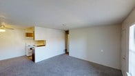 This single-floor residential space offers a straightforward and functional layout designed for comfortable everyday living. The main living area is finished with wall-to-wall carpet in a neutral gray tone, providing a soft and cohesive foundation that complements the light-colored walls throughout the space. The textured ceiling adds a classic touch consistent across the entire unit.

The kitchen is positioned along one wall and features wood-toned cabinetry, a white electric range, and a built-in pass-through counter that visually connects the kitchen to the living area. This open pass-through design promotes a sense of connectivity between the two spaces, making the overall layout feel more open and sociable.

A centrally located doorway leads to the remaining areas of the unit, including what appears to be additional rooms beyond the main living space. Ceiling vents are visible throughout, indicating a central HVAC system for year-round climate control. The overall palette of the space — neutral walls, gray carpet, and warm wood cabinetry — creates a clean, understated aesthetic that offers a versatile canvas for a range of interior styles. The layout is practical and efficiently organized, making good use of available space on a single level.