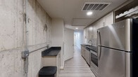 Located at 3410 W Clay St in Richmond, Virginia, this single-floor unit offers a thoughtfully designed layout that maximizes its compact footprint. The galley-style kitchen is a standout feature, showcasing gray shaker cabinetry, dark granite countertops, a subway tile backsplash, and a full suite of stainless steel appliances including a refrigerator, range, and dishwasher. An exposed concrete accent wall adds an industrial character to the kitchen, contrasting beautifully with the clean, modern cabinetry. Light luxury vinyl plank flooring runs throughout the main areas, lending warmth and continuity to the space. A connecting hallway guides you through the unit, leading to a bright open room with a distinctive dark-paneled ceiling and a wide bank of windows fitted with horizontal blinds that allow ample natural light to fill the space. The bathroom features large-format white tile flooring, a white vanity with dark granite countertop, and a generous mirror with overhead lighting. Recessed lighting throughout ensures each area is well-lit. The overall ambiance blends industrial and contemporary design elements, creating a stylish and functional living environment.