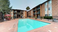 This multi-story residential community showcases a timeless architectural design built with warm, multi-toned brick exteriors and accented by vertical paneling details. The buildings rise several stories, each level featuring private balconies that overlook the well-maintained communal grounds below.

At the heart of the outdoor space is a generously sized swimming pool enclosed by a decorative wrought-iron fence, reserved exclusively for residents and their guests. The pool area features a brick-paved surround, entry handrails, and is complemented by potted tropical plants and seating areas shaded by red umbrellas, creating an inviting recreational retreat.

Adjacent to the pool, a landscaped courtyard provides additional outdoor living space, complete with artificial turf accented by decorative boulders, outdoor picnic tables, and charcoal BBQ grills — ideal for casual gatherings and al fresco dining. Mature pine and deciduous trees are scattered throughout the grounds, providing natural shade and greenery that enhance the community's overall ambiance.

The exterior walkways and building entries are clean and accessible, with well-maintained landscaping including rock beds and shrubs lining the building perimeters. The combination of classic brick construction, thoughtful amenities, and lush communal spaces creates a comfortable and inviting residential environment.