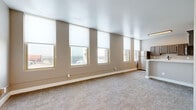 This single-floor unit offers a thoughtfully designed layout with a seamless flow between the kitchen, living, and bedroom areas. The kitchen is a standout feature, equipped with stainless steel appliances including a refrigerator, electric range, over-the-range microwave, and dishwasher. Gray shaker-style cabinetry pairs elegantly with crisp white countertops, while light wood-look flooring grounds the space with warmth and practicality. A peninsula counter provides a convenient transition between the kitchen and the carpeted living area. The expansive main living space is lined with a row of large windows fitted with roller shades, bathing the room in natural light and offering sweeping elevated views of the surrounding cityscape. The bedroom area similarly benefits from multiple windows framing outdoor vistas, complemented by soft carpeting underfoot. Additional interior rooms are accessible through white-paneled doors, with elevated ceilings adding an open, airy quality. The bathroom features a combined tub and shower unit, a single-sink vanity with a mirrored medicine cabinet, and wood-look flooring. Neutral taupe wall tones throughout the unit create a cohesive and calming atmosphere, well-suited for comfortable everyday living.