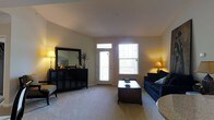 This single-floor space centers around a comfortably sized living room that combines functionality with a warm, inviting ambiance. The room is finished with soft, neutral-toned carpeting that extends throughout, contributing to a cozy feel underfoot. Smooth, light-colored walls and a clean white ceiling help to brighten the interior, while warm accent lighting from strategically placed floor and table lamps adds a welcoming glow.

Natural light enters the living room through a combination of a paneled front door with a transom window above and a large adjacent single-hung window, offering views of the surrounding outdoor environment. These windows together ensure the space remains well-lit throughout the day.

A dark-finished, multi-door storage cabinet along one wall serves as a prominent feature, topped with decorative accessories and flanked by a large framed mirror that visually expands the room. A deep navy-blue sofa with teal accent pillows anchors the seating area, paired with a dark wood rectangular coffee table at the center. The overall design maintains a straightforward, functional aesthetic with neutral tones complemented by darker furniture accents.