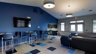 This single-floor community space is thoughtfully designed to serve as both a social hub and a functional gathering area. The room makes a striking visual impact with its bold cobalt blue accent walls contrasted against neutral gray tones, creating an energetic yet welcoming atmosphere.

The flooring features a distinctive patchwork carpet tile design in shades of blue, gray, and beige, adding a dynamic visual element that ties the color palette together. Overhead, contemporary drum-shade pendant lights provide warm, even illumination throughout the space.

Seating is arranged to accommodate a variety of uses. High-top bar tables paired with modern chairs offer a casual spot for group conversation or individual work, while a cluster of upholstered lounge chairs provides a more relaxed seating option. This flexible layout encourages both focused activity and informal gathering.

A kitchenette area is integrated along one wall, featuring dark cabinetry, a countertop workspace, and a sink — offering convenient access to refreshments. Windows along the perimeter allow natural light to filter into the space, enhancing the open and airy feel.

A wall-mounted flat-screen television anchors one end of the room, making the space well-suited for presentations, entertainment, or group viewing. The vaulted ceiling adds a sense of vertical openness, further elevating the overall spaciousness of this well-appointed communal area.