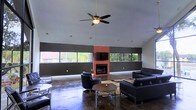 This single-story space offers an open, functional layout across multiple interconnected areas. The centerpiece is a generous lounge room characterized by soaring vaulted ceilings fitted with a ceiling fan, providing a sense of openness and airflow. A striking red brick fireplace serves as the room's focal point, flanked by a wall-mounted television, and is surrounded by a seating arrangement of dark leather chairs and sofas, creating a comfortable gathering environment. Wraparound horizontal windows line the upper portion of the walls, drawing in abundant natural light and offering views of the surrounding greenery. The polished concrete floors extend throughout the space, lending an industrial yet refined aesthetic. Adjacent areas include additional rooms visible from the floor plan, with smaller enclosed spaces likely serving as utility, storage, or support rooms. The overall layout follows an irregular L-shaped configuration, maximizing usable floor area while maintaining a cohesive design language. The combination of high ceilings, natural light, exposed brick, and durable finishes gives the space a modern, versatile character well-suited for communal or commercial use.