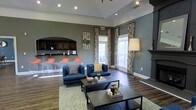 This well-appointed single-floor space combines comfort and style in a thoughtfully designed layout. The main lounge area serves as the focal point, featuring rich hardwood-style flooring, a tray ceiling with recessed lighting, and bold gray walls that create a warm, contemporary ambiance. Navy blue seating is arranged around a pair of sleek dark coffee tables atop a patterned area rug, offering a comfortable and inviting gathering space. Large windows dressed in decorative floral curtains flood the room with natural light, enhancing the open and airy feel of the interior. To one side, a bar counter area is outfitted with dark cabinetry and a row of vibrant orange bar stools, adding a lively pop of color to the space. A classic fireplace mantel anchors one corner of the room, contributing a touch of traditional elegance. Crown molding and white baseboards provide clean architectural definition throughout. The overall aesthetic blends modern furnishings with classic design elements, resulting in a space that is both functional and visually striking — well-suited for socializing, relaxing, or hosting gatherings.