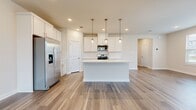 This well-designed two-story home offers five bedrooms and two bathrooms across a thoughtfully laid-out floor plan. The main level features an open-concept kitchen and living area anchored by a striking kitchen island with a built-in sink and crisp white quartz countertops. White shaker cabinetry, a subway tile backsplash, stainless steel appliances — including a French door refrigerator, gas range, and microwave — and elegant gold pendant lighting complete the kitchen. Light wood-look luxury vinyl plank flooring runs throughout the main level, complementing the neutral, warm color palette. Large windows and a rear entry door flood the living area with natural light and provide outdoor views. A clean, well-lit hallway connects the main-level rooms, leading to additional spaces with easy flow. The upper floor houses the majority of the bedrooms, including a spacious primary suite. Bedrooms feature plush carpet, ceiling fans, and windows that invite natural light. The bathrooms are finished with wood-look LVP flooring, white vanity cabinetry, and a glass-enclosed tile shower. Two closets provide ample storage throughout the home. The overall ambiance is clean, bright, and move-in ready, reflecting a modern aesthetic with neutral tones and quality finishes.