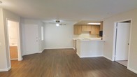 This single-floor residence offers a clean, open-concept layout that seamlessly connects the living area and kitchen into one cohesive space. The living room is finished with rich, dark wood-tone vinyl plank flooring that extends throughout, adding warmth and visual continuity. A ceiling fan with an integrated light fixture centers the room, providing both comfort and illumination. Natural light filters in through a window near the entry door, keeping the space bright and welcoming.

The kitchen is open to the living area via a raised peninsula counter with a light-toned surface, ideal for casual dining or meal prep. Warm honey-toned wood cabinetry lines the upper and lower sections of the kitchen, offering ample storage space. A sink is positioned beneath a skylight or overhead fixture that brightens the work area. A dishwasher is integrated into the cabinetry, and a black refrigerator anchors one end of the kitchen. The walls throughout are painted in a neutral white, creating a clean backdrop that complements the flooring and cabinetry. The overall ambiance is functional and inviting, with a straightforward, practical design that makes efficient use of the available space.
