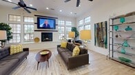 Located at 30685 FM2978 in Magnolia, Texas, this single-story community facility is designed with both comfort and functionality in mind. The space showcases light hardwood flooring throughout, crisp white walls, and soaring ceilings that create an open, airy atmosphere.

The central lounge area serves as the heart of the space, featuring a tufted charcoal sofa, ceiling fan, recessed lighting, and a wall-mounted flat-screen TV above a built-in gas fireplace. An abundance of multi-pane windows floods the room with natural light and provides views of the surrounding landscape. Elegant table lamps and curated artwork add warmth to the contemporary décor.

The kitchen is a standout feature, highlighted by a large granite island with bar seating, white shaker cabinetry, a classic subway tile backsplash, stainless steel appliances, and striking geometric pendant lights overhead. A dedicated coffee and beverage station is conveniently positioned nearby.

A private office or workroom offers built-in shelving with arched detailing, a granite-topped wall desk, and a pendant light, creating a productive and polished workspace. An additional room with glass-paned French doors appears to function as a fitness or multipurpose area. Arched entryways connect the various zones, lending architectural character throughout this thoughtfully designed community space.