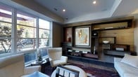Located at 1-3 Mill River Lane in Ardsley, New York, this single-story space presents a thoughtfully designed interior that blends contemporary style with warm, inviting elements. The lounge area serves as a central highlight, showcasing a substantial built-in wall unit crafted from rich, dark wood. This feature wall incorporates open shelving, display niches, and decorative accents, including curated artwork, ceramic vases, and carefully arranged objects that contribute to a gallery-like aesthetic.

Comfortable, upholstered seating in neutral cream tones is arranged to encourage conversation and relaxation, complemented by a richly patterned area rug that anchors the space with deep, earthy tones. Recessed ceiling lighting provides a warm, even illumination throughout the room, enhancing the sophisticated atmosphere.

Large windows along one wall admit generous natural light, offering views of the surrounding greenery and bringing an organic connection to the outdoors. The combination of warm wall tones, dark wood finishes, and layered lighting creates a cohesive, refined ambiance. Overall, the space conveys a polished, contemporary character suitable for comfortable gathering and leisurely use.