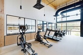 This 540-square-foot fitness facility exemplifies modern design and functionality. The space boasts impressively high ceilings, accentuated by exposed black HVAC ducts that add an industrial aesthetic. Large windows line the walls, flooding the area with natural light and offering views of the surrounding landscape. The room is equipped with a variety of exercise machines, including treadmills, ellipticals, and stationary bikes, strategically placed to maximize space utilization. A full-length mirror spans one wall, allowing users to monitor their form during workouts. The neutral color palette, featuring white walls and light flooring, creates a clean, open atmosphere. Blue accents in the seating provide subtle pops of color. The facility's layout promotes easy navigation and access to equipment, while glass partitions separate different workout zones, maintaining an open feel while defining distinct areas for various exercise routines.