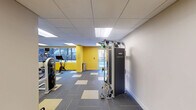 Located at 116 W 9th St in Wilmington, Delaware, this fitness center offers a generously sized, single-floor exercise room designed to accommodate a wide range of workout needs. The space is defined by its bold yellow accent walls, which contrast sharply with neutral white and grey tones throughout, creating an energetic and motivating atmosphere.

The exercise room is outfitted with a comprehensive selection of fitness equipment, including treadmills, stationary bikes, a rowing machine, and multi-station cable machines such as the Technogym Instinct. Free weights, kettlebells, and stability balls are also available, catering to both strength training and functional fitness routines. A dedicated wall-mounted accessory station adds further versatility to the workout options.

Large mirrors line a significant portion of the walls, enhancing the sense of openness and allowing users to monitor their form during exercise. The drop ceiling features recessed fluorescent panel lighting that evenly illuminates the entire floor, while windows along one wall allow natural light to supplement the interior lighting.

The flooring consists of a patterned modular carpet tile in grey and gold tones, reinforcing the space's dynamic color scheme. The layout is open and thoughtfully organized, ensuring ease of movement between equipment stations. This fitness center provides a functional and well-maintained environment suited for a comprehensive workout experience.