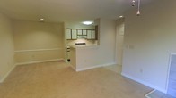 This single-floor space features an open-concept design that seamlessly connects the living area to the kitchen. The layout is anchored by wall-to-wall beige carpeting throughout the main living space, contributing to a warm and understated atmosphere. Neutral cream and white wall tones keep the interior feeling bright and cohesive.

The kitchen is positioned behind a half-wall breakfast bar, creating a natural division between the cooking and living areas while maintaining visual openness. It is equipped with a white electric range, an overhead range hood, and a combination of white and wood-toned upper cabinetry. Recessed lighting and a flush-mount ceiling fixture provide functional illumination in the kitchen area.

Recessed ceiling lights are also present in the main living space, supplementing the overall brightness. A standard interior door off the living area leads to an additional room or closet space. White baseboards run along the perimeter of the space, adding a clean, finished look. Overall, the space presents a practical and straightforward layout suited for everyday living.