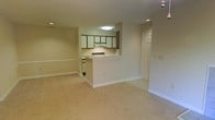 This single-floor space features an open-concept design that seamlessly connects the living area to the kitchen. The layout is anchored by wall-to-wall beige carpeting throughout the main living space, contributing to a warm and understated atmosphere. Neutral cream and white wall tones keep the interior feeling bright and cohesive.

The kitchen is positioned behind a half-wall breakfast bar, creating a natural division between the cooking and living areas while maintaining visual openness. It is equipped with a white electric range, an overhead range hood, and a combination of white and wood-toned upper cabinetry. Recessed lighting and a flush-mount ceiling fixture provide functional illumination in the kitchen area.

Recessed ceiling lights are also present in the main living space, supplementing the overall brightness. A standard interior door off the living area leads to an additional room or closet space. White baseboards run along the perimeter of the space, adding a clean, finished look. Overall, the space presents a practical and straightforward layout suited for everyday living.