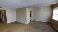 This two-story residential space offers a straightforward and functional layout designed for comfortable everyday living. The main living area is finished with wall-to-wall carpeting in a warm neutral tone, lending a cozy feel to the space. A textured ceiling adds subtle dimension overhead, while clean white baseboards frame the room throughout.

A partial interior wall divides the open-plan area, creating a natural separation between the living and dining zones. The dining area is highlighted by a multi-bulb chandelier that provides warm ambient lighting. Adjacent to the dining space, tile flooring transitions into what appears to be a kitchen or utility corridor, providing a practical and easy-to-clean surface in higher-traffic zones.

A dedicated laundry closet is conveniently tucked into the layout, visible through an open doorway along the dividing wall — a practical feature that adds significant everyday convenience. Neutral beige walls throughout create a clean, versatile backdrop suitable for a variety of interior styles.

HVAC vents are thoughtfully positioned in the ceiling and walls to ensure consistent climate control. The overall design is simple and understated, prioritizing utility and livability. With two floors, the space offers ample room to separate living and sleeping areas, making it well-suited for a range of occupants.