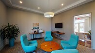 Located at 535 Blackrock Dr in Blacksburg, Virginia, this single-floor community space offers a polished and inviting environment designed for comfort and functionality. The space is anchored by a stylish lounge area featuring a cluster of vibrant teal swivel chairs arranged around a warm wood-toned circular coffee table, creating an ideal setting for relaxed conversation or casual gatherings. Patterned carpeting with flowing motifs adds visual interest underfoot throughout the main area. A wall-mounted flat-screen television is paired with a sleek console table, providing both entertainment and aesthetic appeal. A dedicated workstation area includes a computer and printer, catering to practical needs. Lush potted plants are thoughtfully placed throughout, adding a refreshing natural element to the interior. Large windows flood the space with abundant natural light, enhancing the open and airy atmosphere. Tray ceilings with recessed lighting and a statement drum pendant light contribute to the refined architectural character of the room. Dark wood-framed glass doors connect the lounge to a bright entry vestibule and adjoining corridors, ensuring a seamless and welcoming flow throughout the entire space.