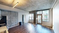 Located on the seventh floor at 1040 G St in the heart of downtown San Diego, this single-floor unit showcases a compelling industrial-modern aesthetic throughout. Exposed concrete ceilings are a defining architectural feature, lending a raw, urban character that is complemented by rich hardwood flooring found throughout the space.

The open living area is bathed in natural light from large windows and a glass-paneled door that opens to city views of the surrounding downtown skyline. Concrete columns and accent walls add structural texture and visual interest, reinforcing the industrial design language.

The kitchen is well-appointed with stainless steel appliances — including a refrigerator, range, microwave, and dishwasher — paired with two-tone cabinetry featuring white upper cabinets and dark wood lower cabinets, finished with granite-style countertops. Linear overhead lighting illuminates the space efficiently.

The bedroom is a bright, airy room with large windows overlooking the cityscape and exposed concrete ceiling details that carry through from the main living area. A closet with louvered bifold doors provides convenient storage, situated adjacent to the bathroom.

The bathroom features a dark wood vanity with a granite countertop, an integrated sink, a large mirror, and hardwood-style flooring, offering a clean and cohesive finish consistent with the rest of the unit.