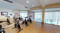 This single-floor indoor cycling studio is thoughtfully designed to support a focused and energizing workout experience. The room is anchored by a row of professional-grade spin bikes arranged on smooth hardwood flooring, providing a clean and durable surface well-suited for high-intensity training sessions. Floor-to-ceiling mirrors line one wall, visually expanding the space and allowing participants to monitor their form throughout a ride. Large windows along the opposite wall invite abundant natural light, creating an open and uplifting atmosphere while offering views to the exterior. Ceiling fans are suspended overhead, supporting ventilation and comfort during intense workouts. A textured accent wall in warm tones adds a visual focal point and a sense of depth to the room. Recessed lighting in the clean white ceiling supplements natural light during evening sessions. The overall design balances functionality with a modern, polished aesthetic — a welcoming environment for both casual riders and dedicated fitness enthusiasts. The space is well-ventilated, open in feel, and efficiently laid out to accommodate a group cycling class comfortably.