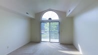 This single-floor space makes a strong architectural impression through its vaulted ceilings and a distinctive arched transom window that crowns a set of full-height sliding glass doors. The combination creates a dramatic focal point, drawing the eye upward while flooding the interior with an abundance of natural light throughout the day. The sliding glass doors provide direct access to an outdoor area framed by lush greenery, seamlessly connecting the interior to the natural surroundings outside. The main living area features neutral-toned carpeting that complements the light, airy color palette of the walls and ceiling. Ceiling vents are thoughtfully integrated into the upper portions of the vaulted ceiling, maintaining the clean, uncluttered aesthetic of the space. The overall ambiance is bright, open, and tranquil, with the greenery visible through the expansive glazing lending the interior a calm, nature-connected feel. The architectural detailing of the arched window adds a refined, classic character to what is otherwise a straightforward and functional layout, making this space both visually appealing and practically designed for comfortable everyday living.