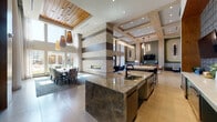 This elegantly designed single-floor communal space presents a seamless blend of modern luxury and warm, natural materials throughout. The interior is defined by dramatic double-height ceilings, with sections clad in warm wood slat paneling and others finished with refined coffered detailing — together creating a visually rich and inviting atmosphere.

At the heart of the space, a striking floor-to-ceiling accent wall constructed of stacked natural stone in alternating light and dark horizontal bands frames a sleek linear fireplace, serving as a focal point for both the lounge and dining areas. The lounge features light hardwood flooring, stylish dark armchairs arranged around a circular wood coffee table, and floor-to-ceiling glass doors that open to an outdoor pool area, flooding the space with natural light and offering seamless indoor-outdoor connectivity.

The dining area accommodates a large group around a long wooden table with upholstered chairs, anchored by a blue vintage-style area rug beneath a cluster of woven pendant lights. Oversized windows frame views of the outdoor pool courtyard.

The kitchen zone is equipped with substantial granite-topped islands featuring undermount sinks, complemented by a bold metallic tile backsplash, green cabinetry, and bar-height seating. Curated artwork and designer pendant lighting complete this polished, contemporary communal retreat.