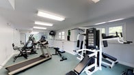 Located at 69 Newfield in Waterbury, this fitness center offers a clean, well-organized workout facility designed to support a full range of exercise routines. The single-floor space is illuminated by rows of overhead fluorescent lighting, ensuring a bright and energizing environment throughout. The interior features a textured ceiling and crisp white walls that contribute to a fresh, open atmosphere.

The gym is equipped with a variety of cardio machines, including treadmills and an elliptical trainer, positioned to allow comfortable spacing between users. A dedicated strength training area includes a multi-function cable and weight machine rated at 950 lbs, alongside a weight bench, providing ample options for resistance training. The flooring features a durable, cushioned surface in a muted green tone, well-suited for high-traffic fitness use.

Natural light filters in through several windows along the wall, offering views of the surrounding greenery and adding a welcoming touch to the space. A wall-mounted television is positioned for easy viewing during workouts. A counter area along one wall provides additional utility for the space. Overall, this fitness center is a practical and well-maintained facility suited for a broad range of fitness needs.