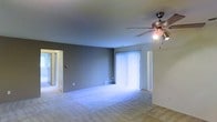 This single-floor space features an open and airy living area that offers a functional, straightforward layout. The room is finished with wall-to-wall carpeting in a neutral tone, providing a soft underfoot feel that complements the understated color palette of the grey-painted walls. A ceiling fan with an integrated light fixture is centrally mounted, offering both overhead illumination and air circulation year-round. A large window dressed with vertical blinds serves as a prominent feature along one wall, allowing natural light to fill the space while providing privacy control. A baseboard heating unit is positioned beneath the window, ensuring comfortable temperatures throughout the cooler months. A doorway visible along the adjacent wall leads to additional rooms, suggesting a connected and accessible floor plan. The overall ambiance is clean and neutral, offering a blank canvas well-suited for a variety of interior arrangements and personal styles. The space is practical in its design, with simple finishes and an uncluttered layout that maximizes usable floor area.
