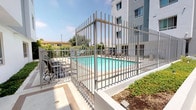 Located at 7147 Woodley Ave in the Van Nuys neighborhood of Los Angeles, this outdoor amenity space is thoughtfully designed to offer residents a well-rounded open-air experience. At the heart of the area is a generously sized swimming pool with clear, blue-tinted water, enclosed by sleek metal security fencing that provides both safety and a modern aesthetic. A tiled pool deck surrounds the water, furnished with multiple lounge chairs and side tables, ideal for relaxing under the Southern California sun.

Adjacent to the pool, a paved courtyard pathway with a two-tone concrete design connects various outdoor zones, creating an organized and visually appealing layout. Raised planters line the walkways, filled with a lush variety of greenery including bamboo, ornamental grasses, fruit trees, and flowering shrubs, contributing to a tranquil, garden-like atmosphere. A dedicated outdoor dining area features a table, chairs, and a shaded umbrella alongside a barbecue grill station, making it well-suited for casual gatherings.

The multi-story building facade, finished in a clean gray stucco, frames the courtyard on multiple sides, with large windows overlooking the outdoor space. The overall design balances functionality with landscaped beauty, offering a welcoming communal retreat.