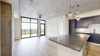 This contemporary 2-bedroom, 2-bathroom unit showcases an industrial-modern aesthetic defined by exposed concrete ceilings, light wood-tone flooring, and crisp white walls throughout. The open-plan kitchen and living area forms the heart of the unit, where floor-to-ceiling black-framed sliding doors flood the space with natural light and provide access to a private balcony with outdoor views. The kitchen is outfitted with dark flat-front cabinetry extending to the ceiling, white quartz countertops, stainless steel appliances, and a curved center island accented by globe pendant lighting.

The living area is generously proportioned and benefits from the same dramatic glazing, with a ceiling fan overhead and an exposed concrete accent wall adding textural contrast. Both bedrooms continue the concrete ceiling detail, each featuring floor-to-ceiling windows, ceiling fans, and access to the balcony. The primary bedroom includes a spacious walk-in closet with built-in wire shelving.

The bathrooms reflect the same refined palette, with a dual-sink vanity featuring dark cabinetry, matte black hardware, floating shelves, and a frameless mirror. A central hallway connects the rooms efficiently, with a built-in open shelving unit providing additional storage near the entry.